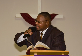 Bishop Anderson in a brown suit, holding a mic, preaching the word of God